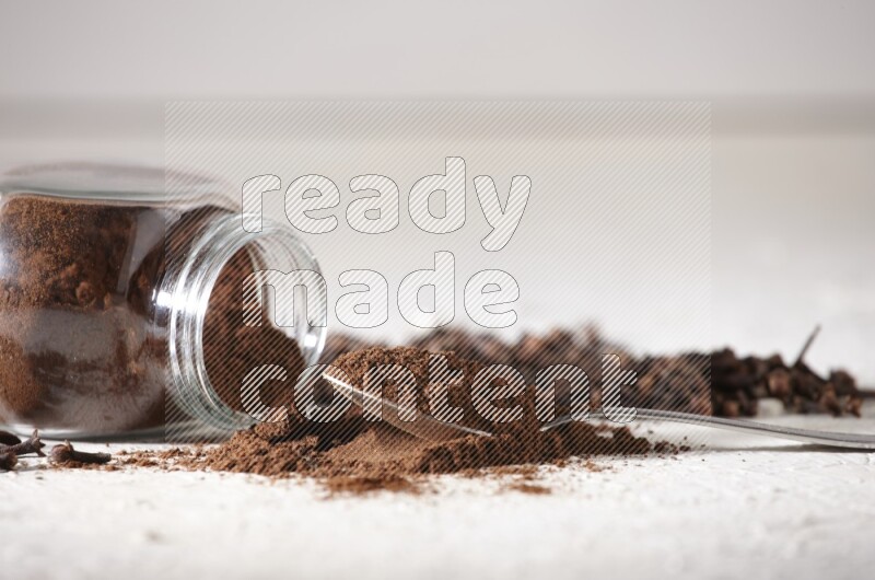 A flipped glass spice jar and a metal spoon full of cloves powder and powder came out of the jar with cloves spread on textured white flooring
