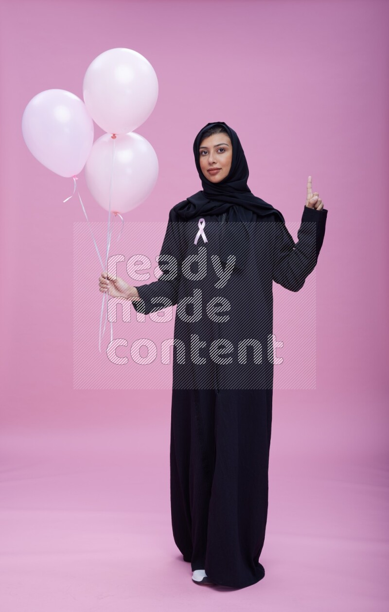 Saudi woman wearing pink ribbon on Abaya standing holding balloons on plain pink background
