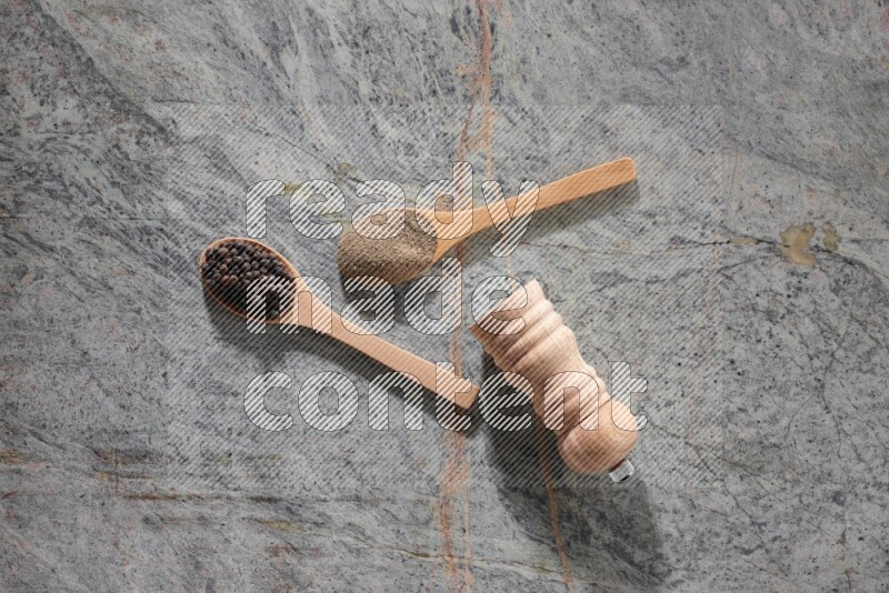 ملعقتان خشبيتان، ملعقة ممتلئة بمسحوق الفلفل الأسود والأخرى ممتلئة بحبات الفلفل الأسود ومطحنة خشبية على خلفية رخام رمادية