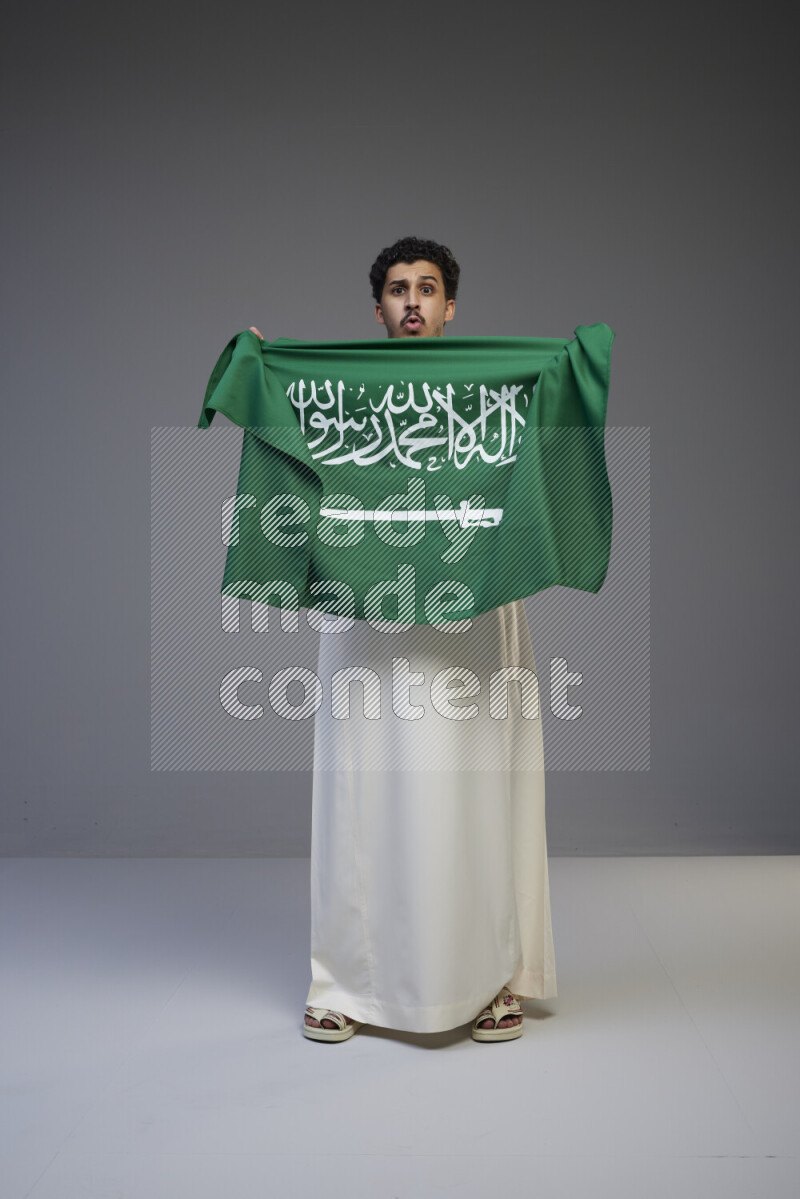 A Saudi man standing wearing thob holding big Saudi flag on gray background