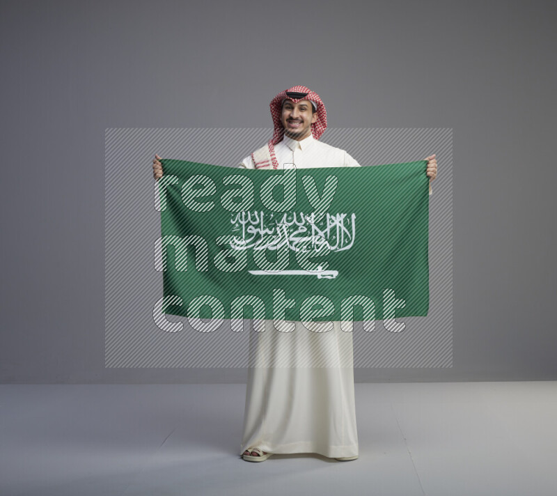 A saudi man standing wearing thob and red shomag holding big saudi flag on gray background