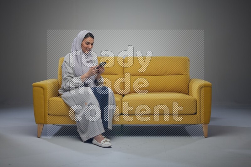 A Saudi woman wearing a light gray Abaya and white head scarf sitting on a yellow sofa and using her phone eye level on a grey background