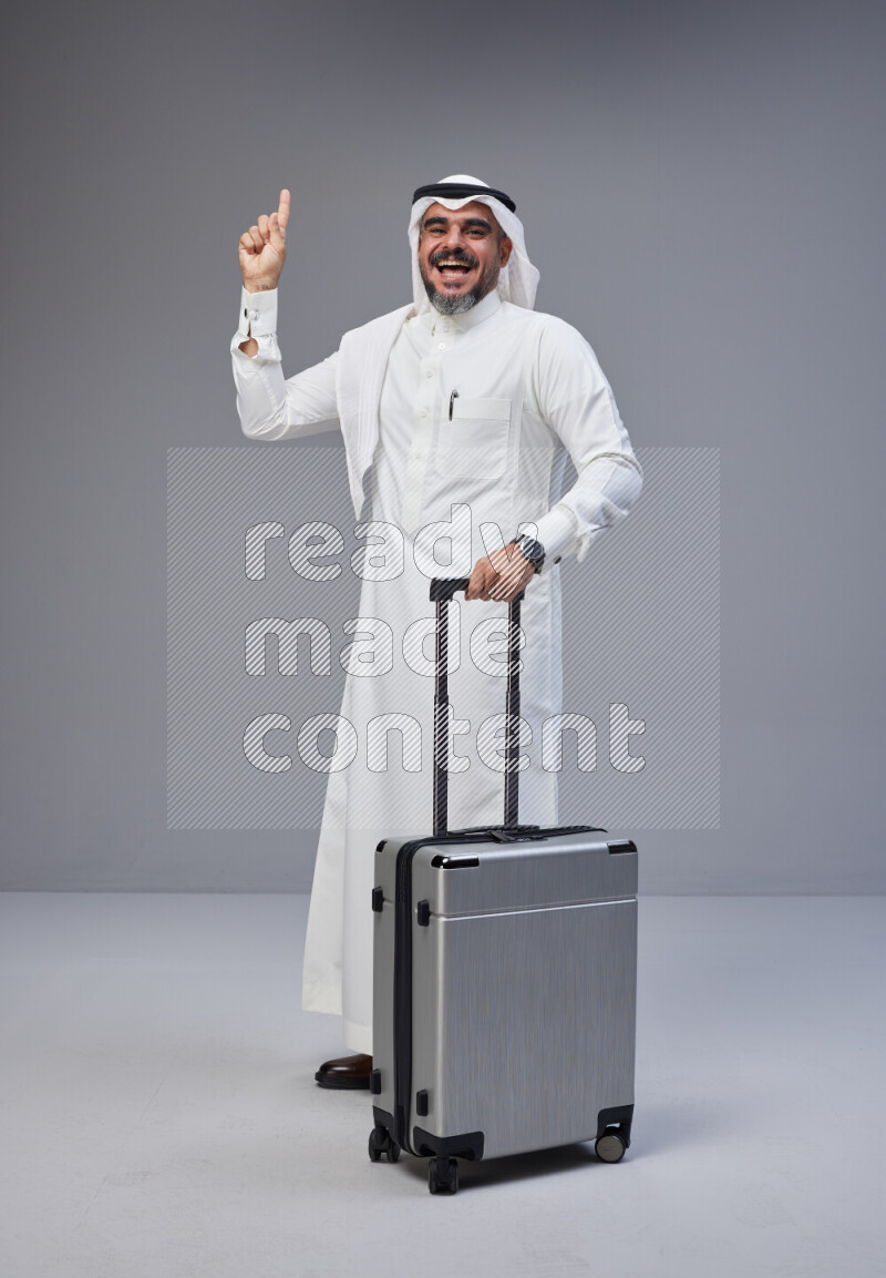 Saudi man wearing Thob and white Shomag standing holding Travel bag on Gray background