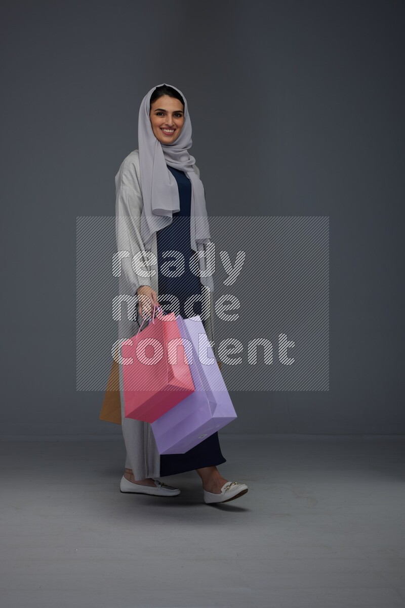 A Saudi woman wearing a light gray Abaya and head scarf standing and holding shopping bags on a grey background