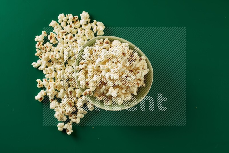 A green ceramic bowl full of popcorn with popcorn beside it on a green background in a top view shot