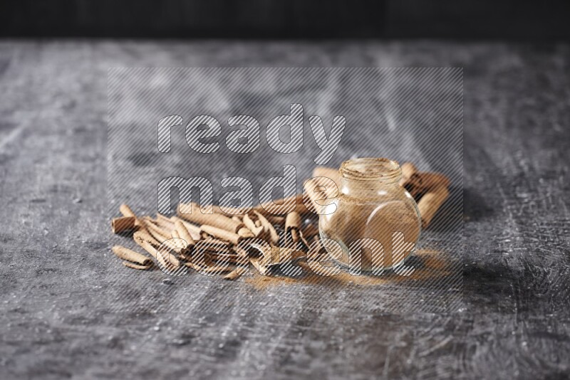 Herbal glass jar and a metal spoon full of cinnamon powder surrounded by cinnamon sticks on textured black background