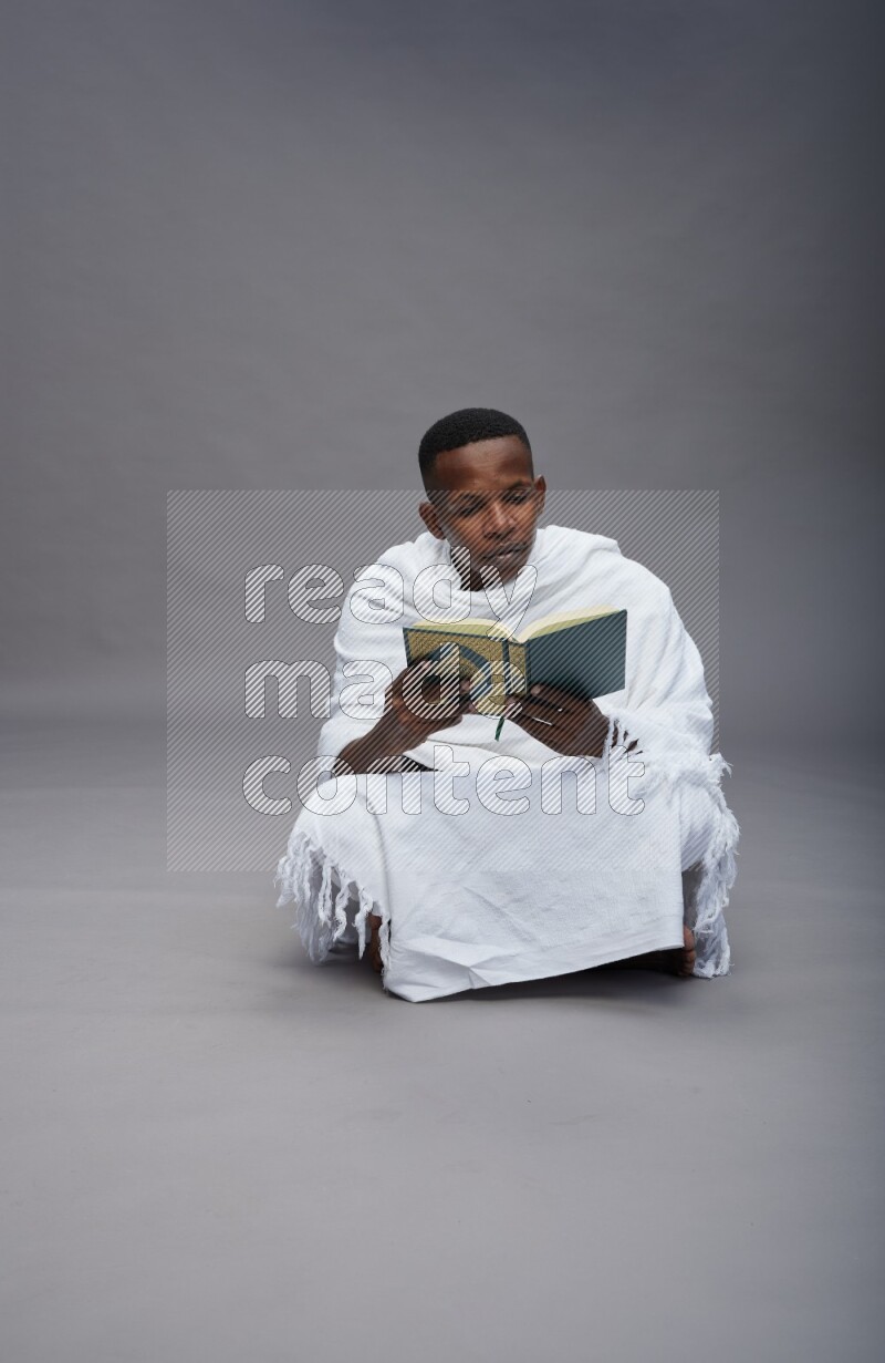 A man wearing Ehram sitting on floor reading quran on gray background