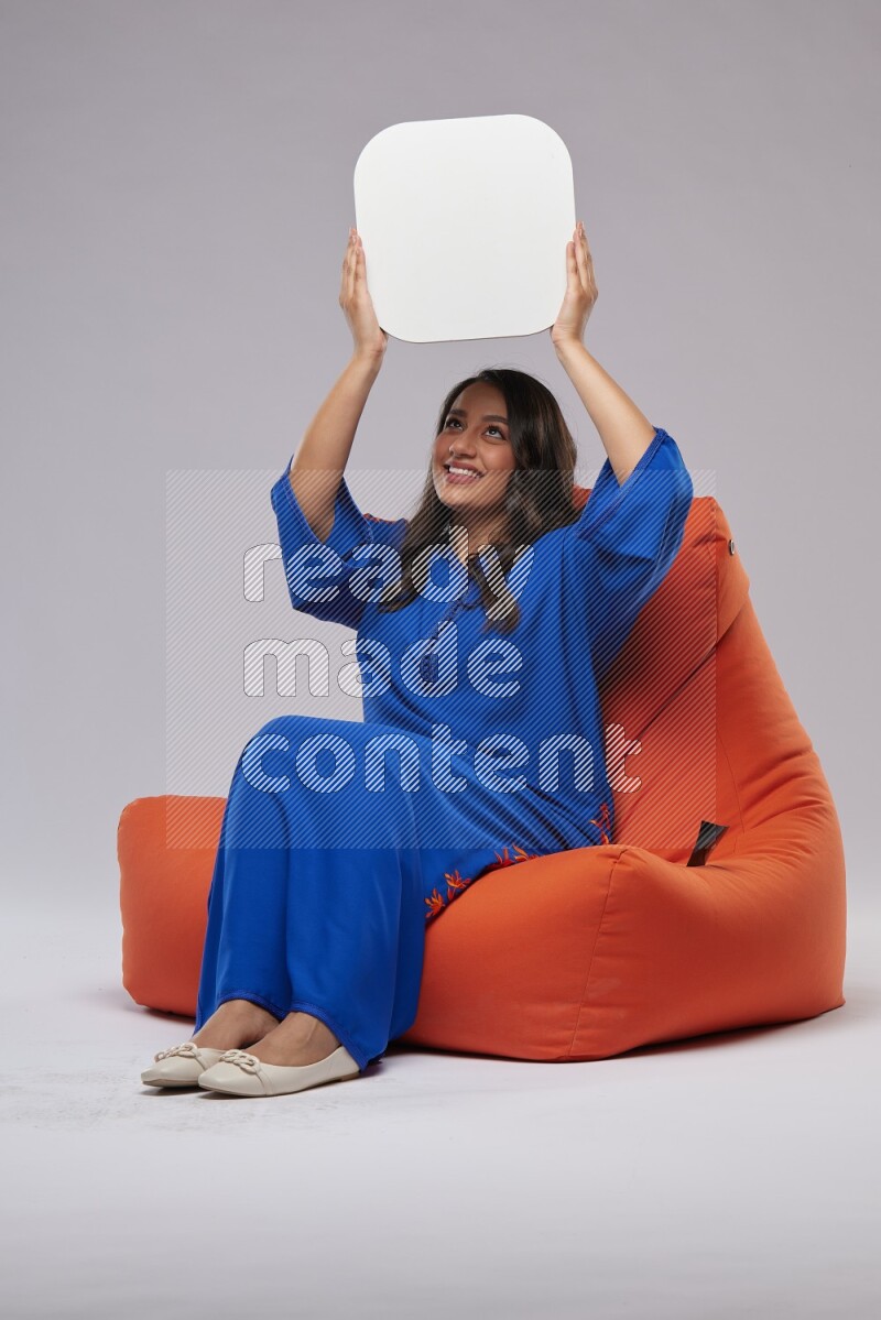 A Woman sitting on an orange beanbag wearing Jalabeya holding a social media sign