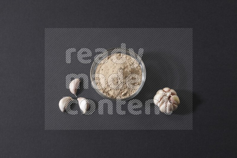 وعاء زجاجي ممتلئ ببودرة الثوم مع بعض فصوص الثوم ورأس ثوم كاملة على خلفية سوداء