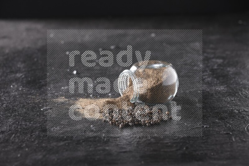 A flipped glass spice jar full of black pepper powder with pepper beads on textured black flooring