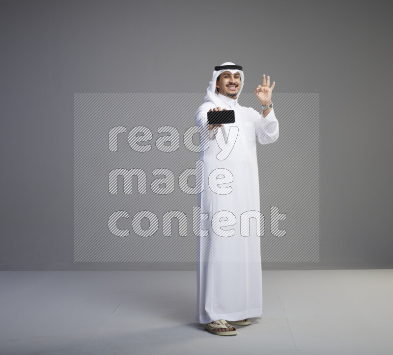 A Saudi man standing wearing thob and white shomag showing phone to camera on gray background