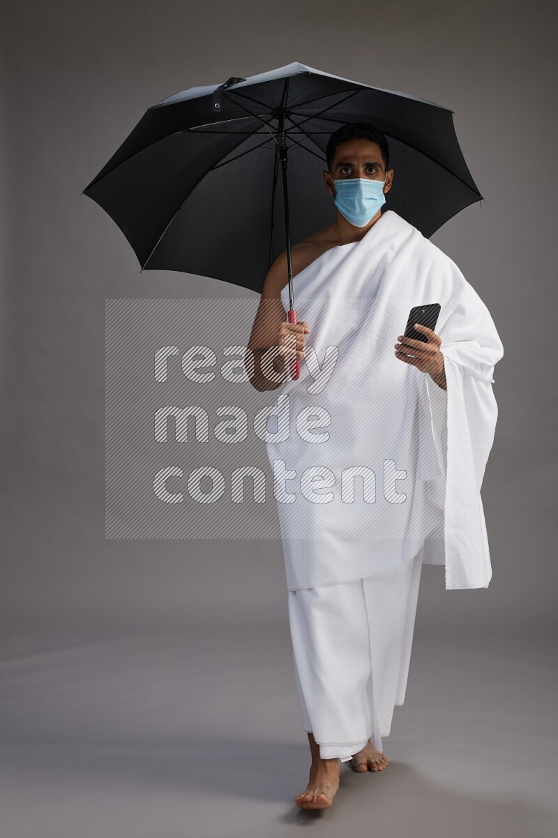 A man wearing Ehram and mask Standing holding umbrella on gray background on gray background