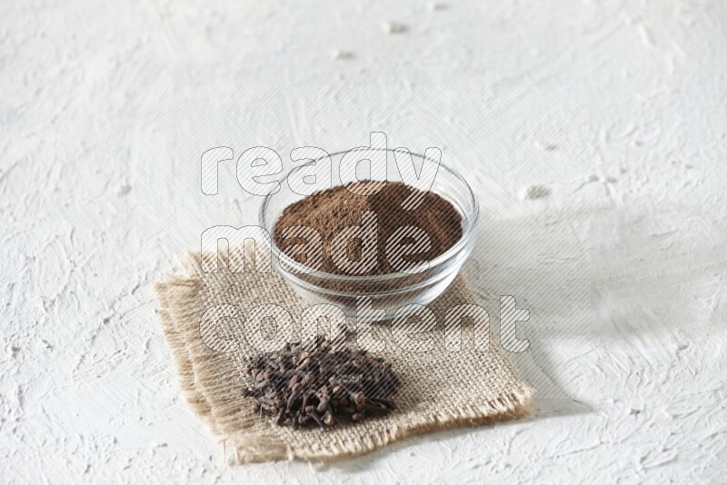 A glass bowl full of cloves powder and cloves grains on a burlap piece on a white flooring
