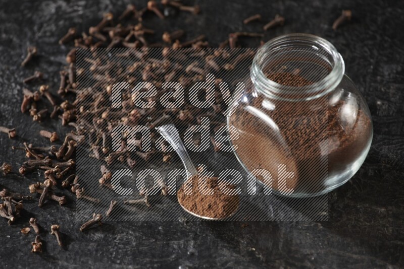 A glass spice jar and a metal spoon full of cloves powder with cloves spread on textured black flooring