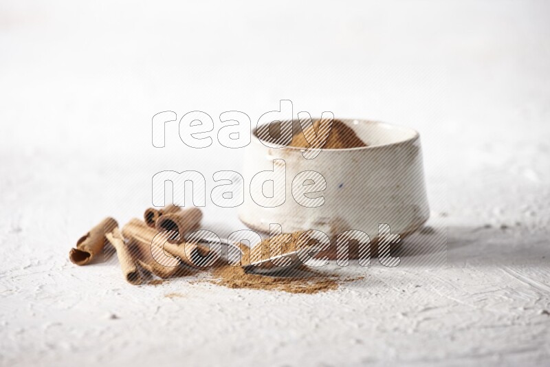 Ceramic beige bowl full of cinnamon powder and a metal spoon with cinnamon sticks next of it on a textured white background