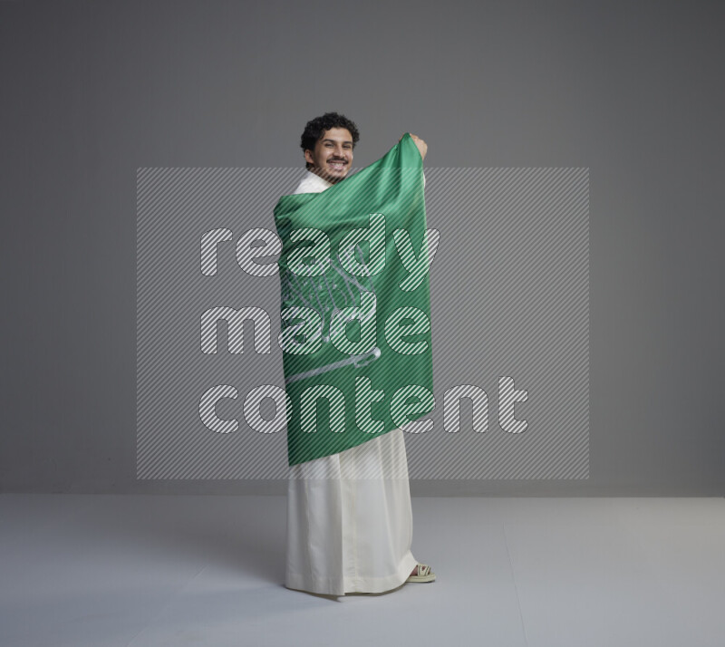 A Saudi man standing wearing thob wrapping big Saudi flag on gray background