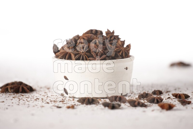 اليانسون النجمي في وعاء أبيض والمزيد من اليانسون متناثر على خلفية بيضاء