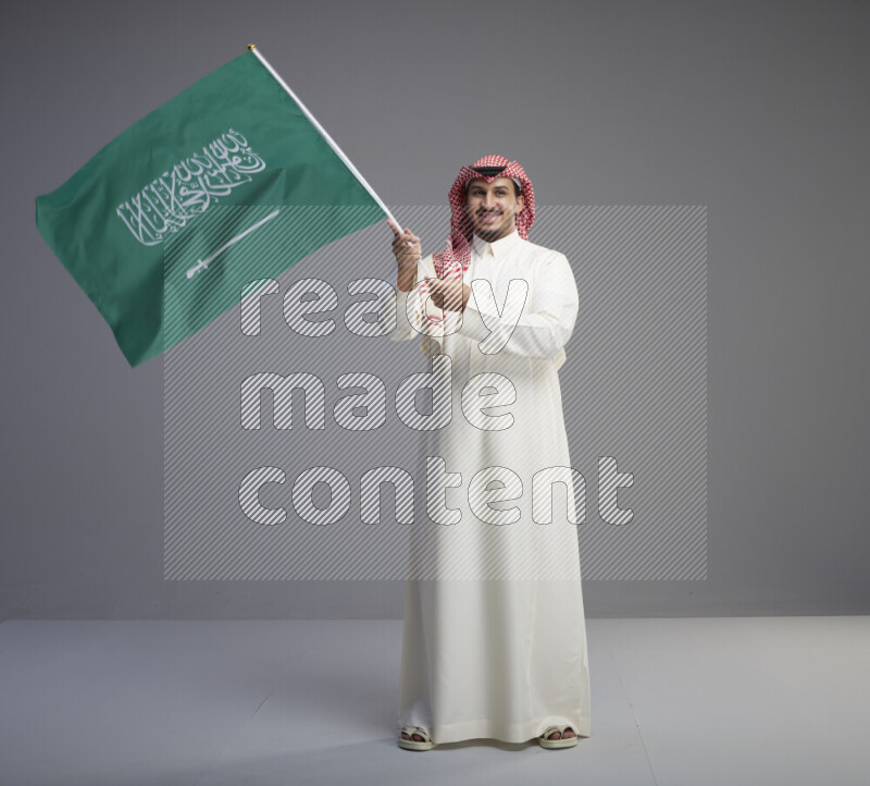 A Saudi man standing wearing thob and red shomag raising big Saudi flag on gray background