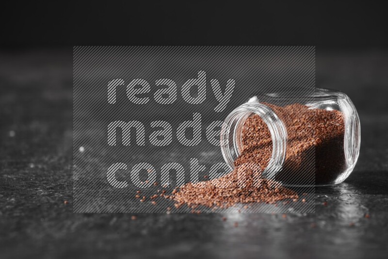 A glass spice jar full of garden cress seeds flipped and seeds spread out a textured black flooring