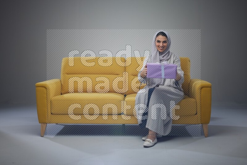 A Saudi woman wearing a light gray Abaya and head scarf sitting on a yellow sofa holding a gift box beside multi color gift boxes eye level on a grey background