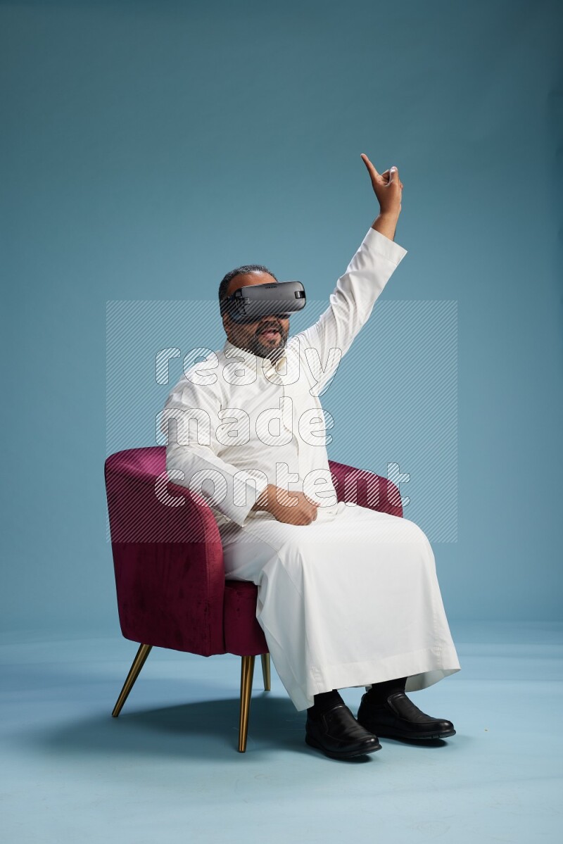 Saudi Man without shimag sitting on chair  gaming with VR on blue background