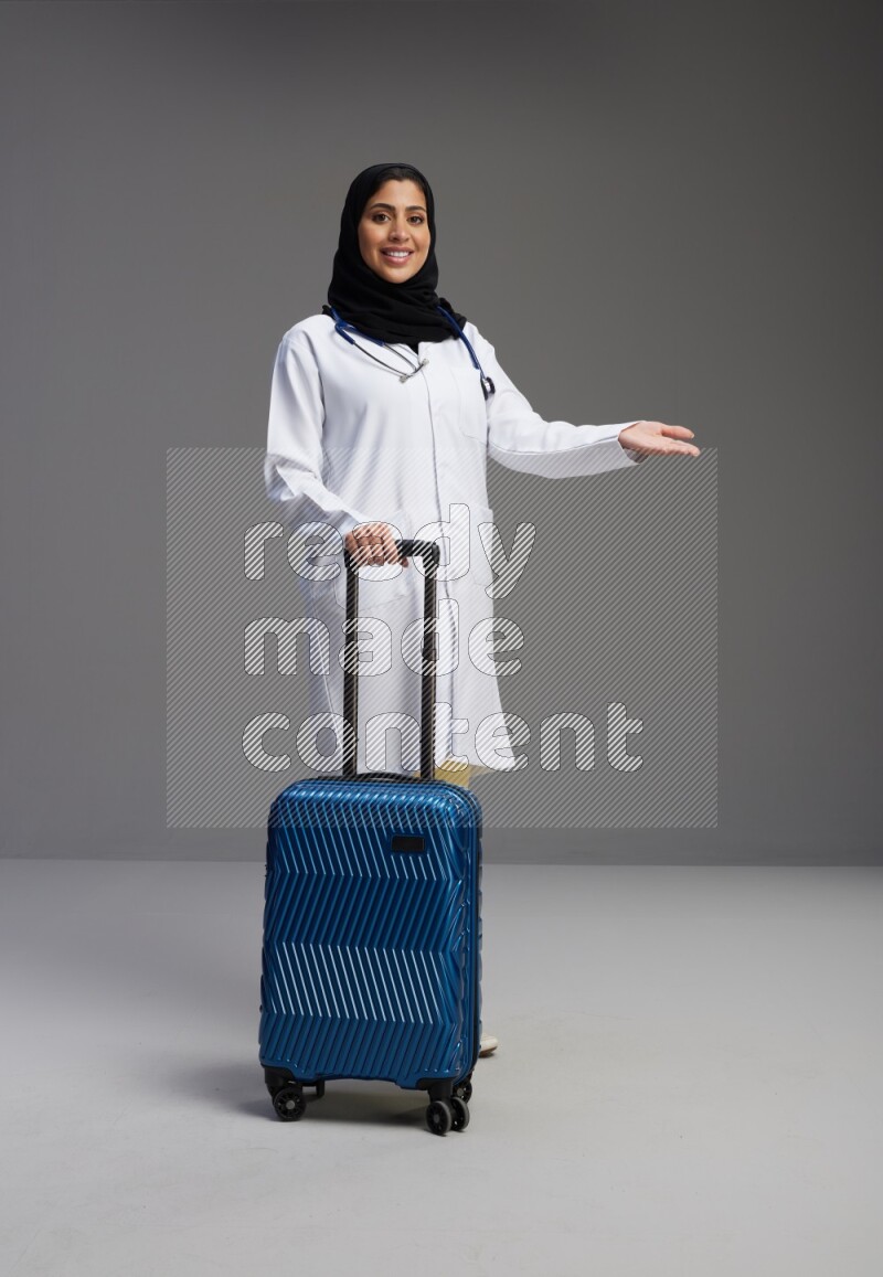 Saudi woman wearing lab coat with stethoscope standing holding Travel bag on Gray background
