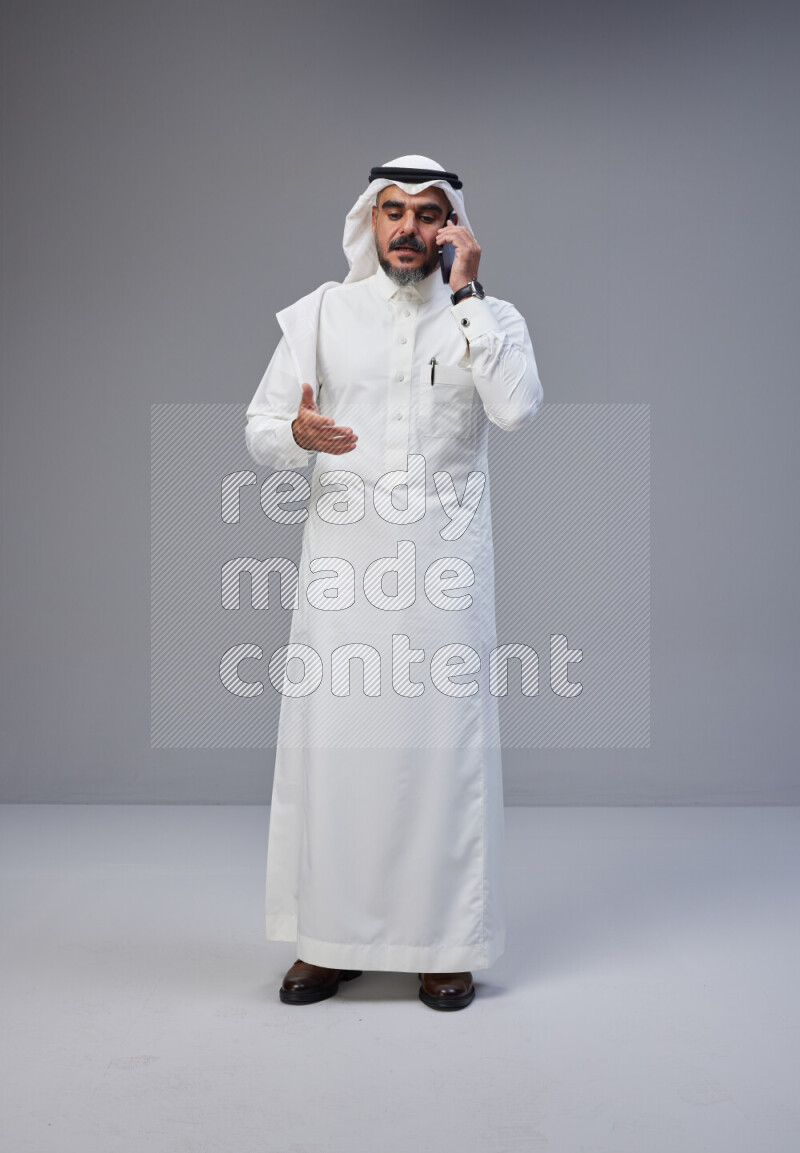 Saudi man Wearing Thob and white Shomag standing talking on phone on Gray background