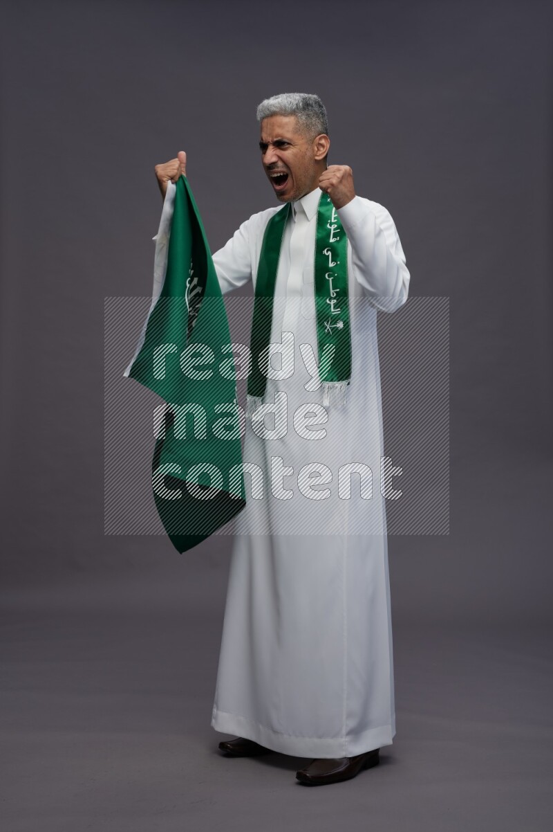 Saudi man wearing thob standing holding Saudi flag on gray background