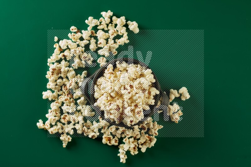 A black ceramic bowl full of popcorn with popcorn beside it on a green background in a top view shot