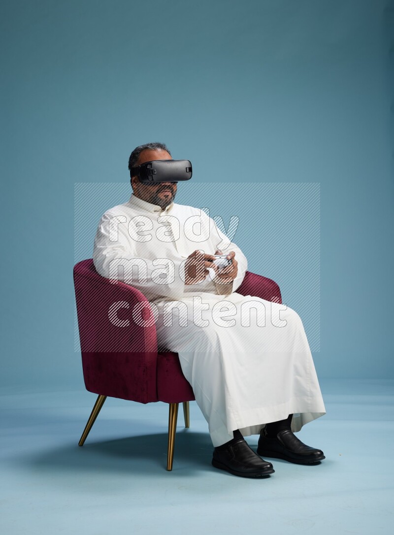 Saudi Man without shimag sitting on chair  gaming with VR and joystick on blue background