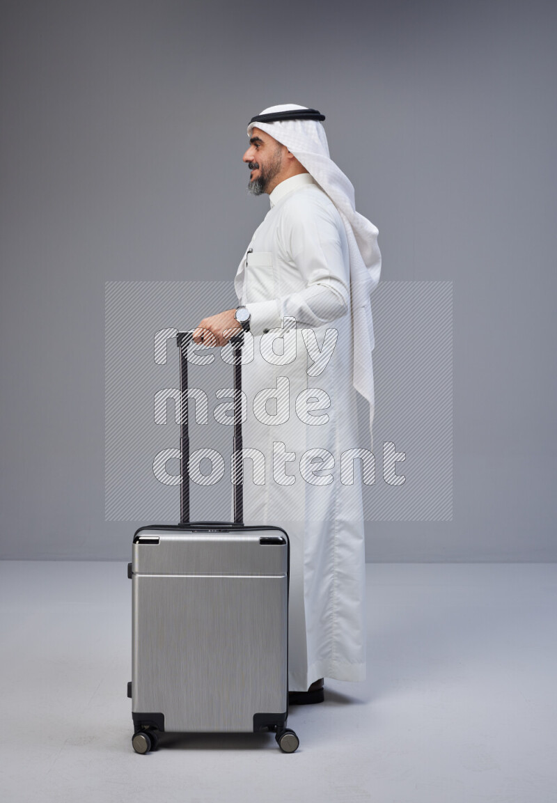 Saudi man wearing Thob and white Shomag standing holding Travel bag on Gray background