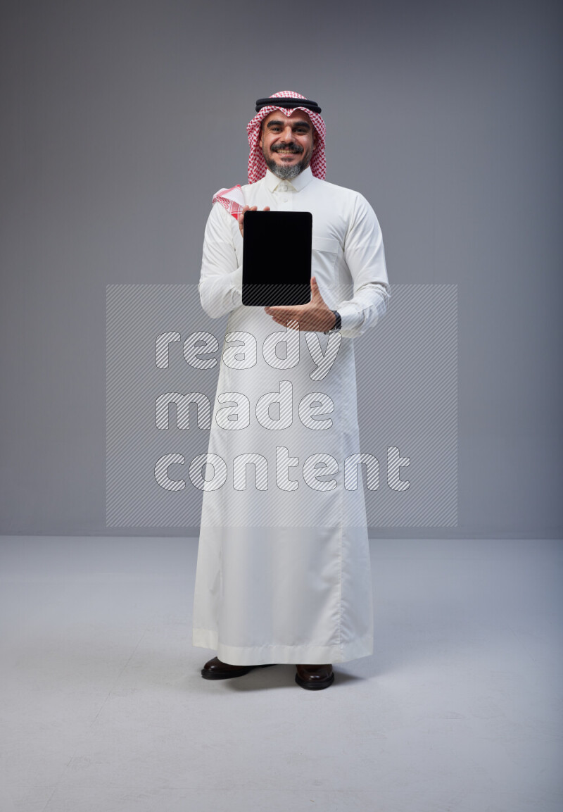 Saudi man Wearing Thob and red Shomag standing showing tablet to camera on Gray background
