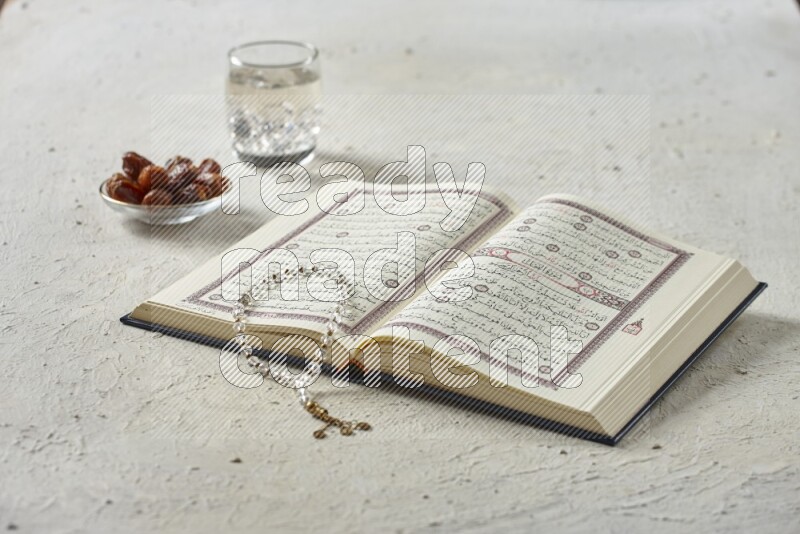 Quran with dates, prayer beads and different drinks all placed on textured white background