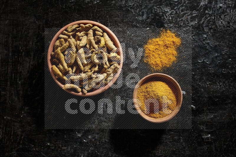 2 wooden bowls, one full of turmeric powder and the other full of dried turmeric whole fingers on a textured black flooring