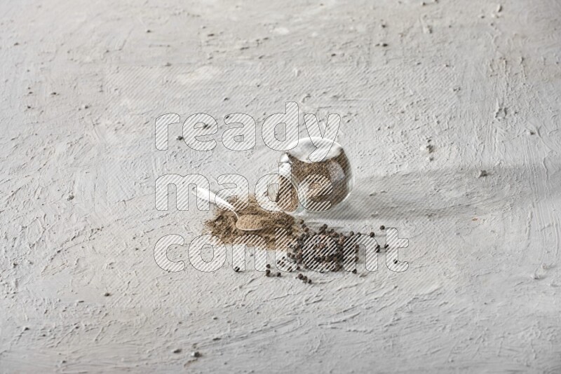 مسحوق الفلفل الأسود في وعاء زجاجي مقلوب وملعقة معدنية وحبات الفلفل علي خلفية بيضاء