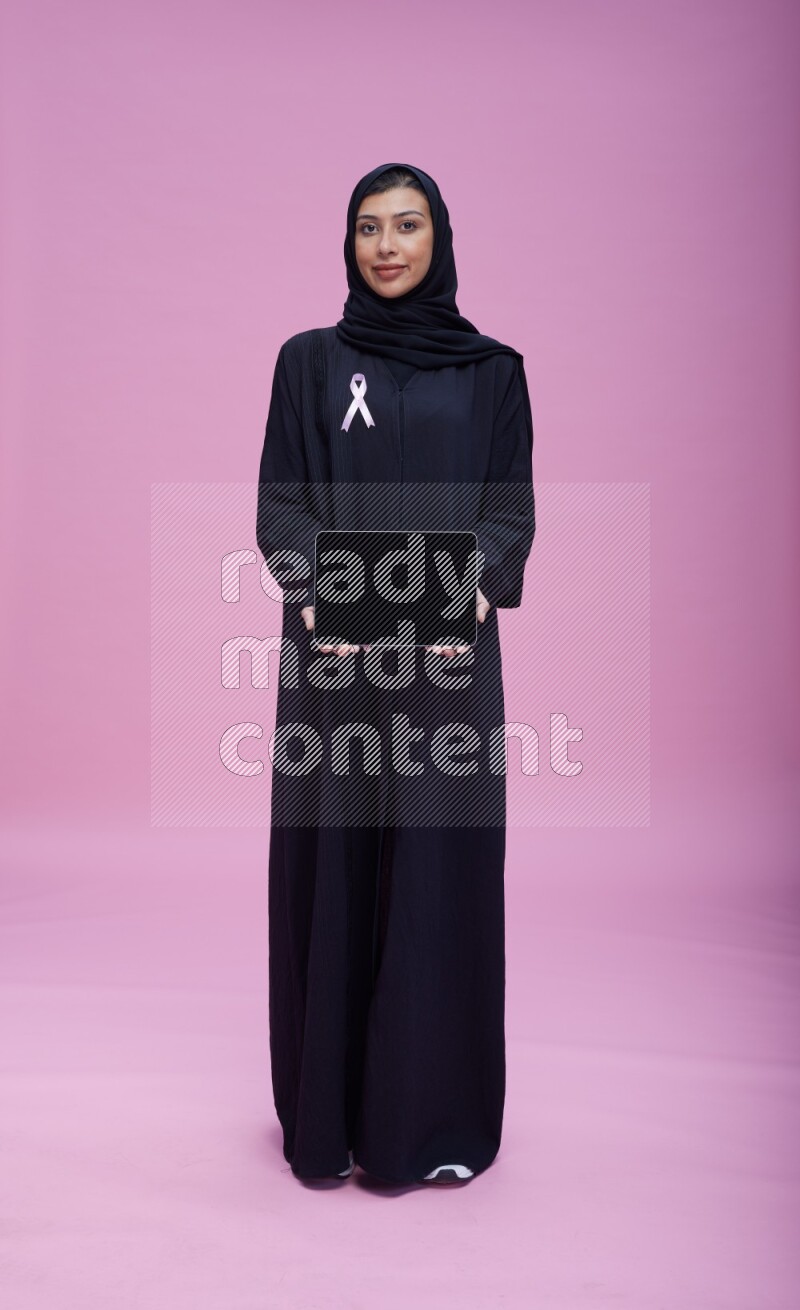 Saudi woman wearing pink ribbon on Abaya standing showing a tablet to camera on pink background