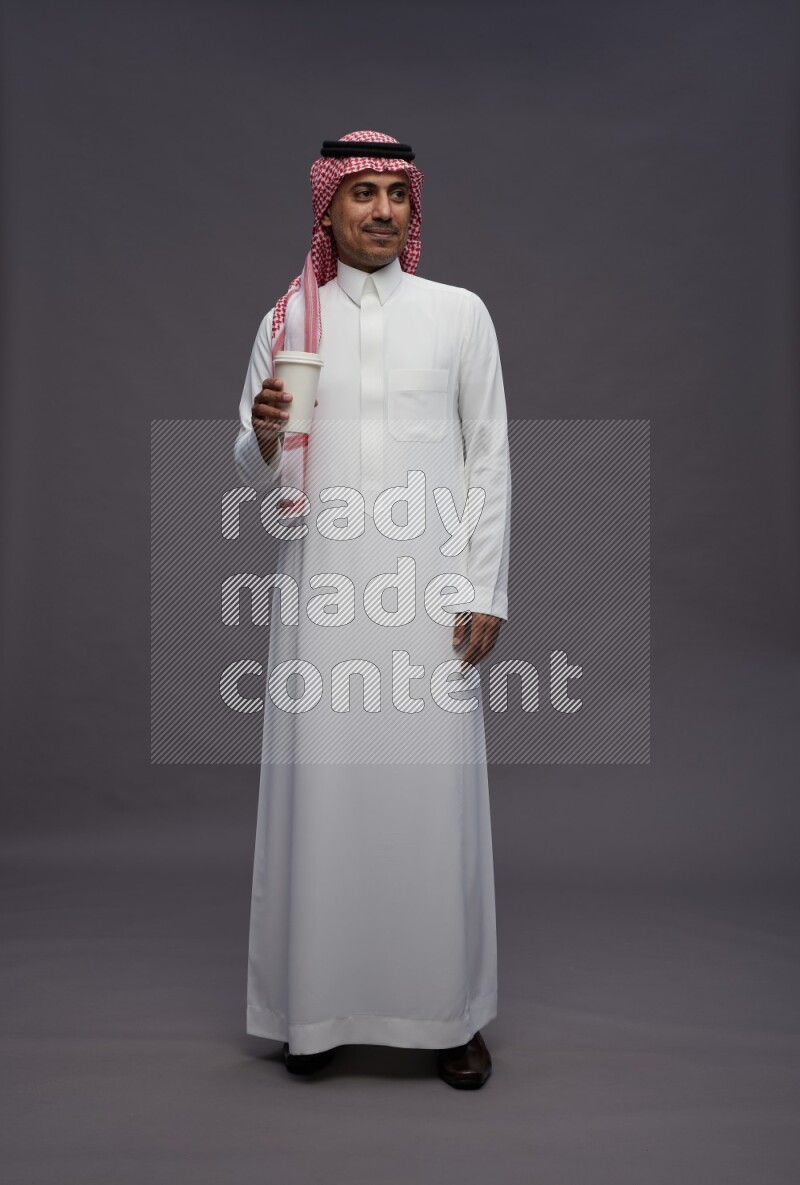 Saudi man wearing thob and shomag standing holding paper cup on gray background