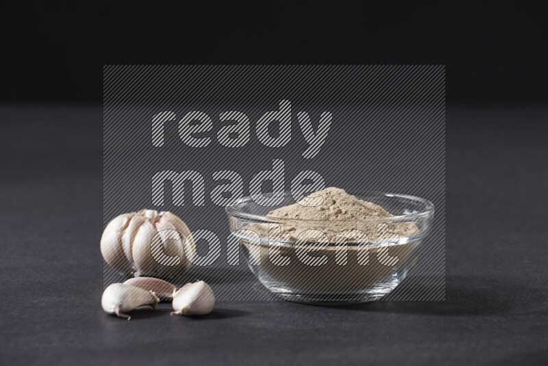 A glass bowl full of garlic powder with garlic bulb and some cloves beside it on a black flooring