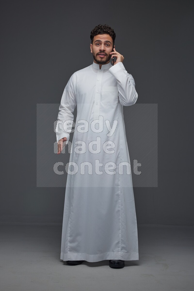 A Saudi man wearing Thobe and talking in the phone while standing and making different hand gestures eye level on a gray background