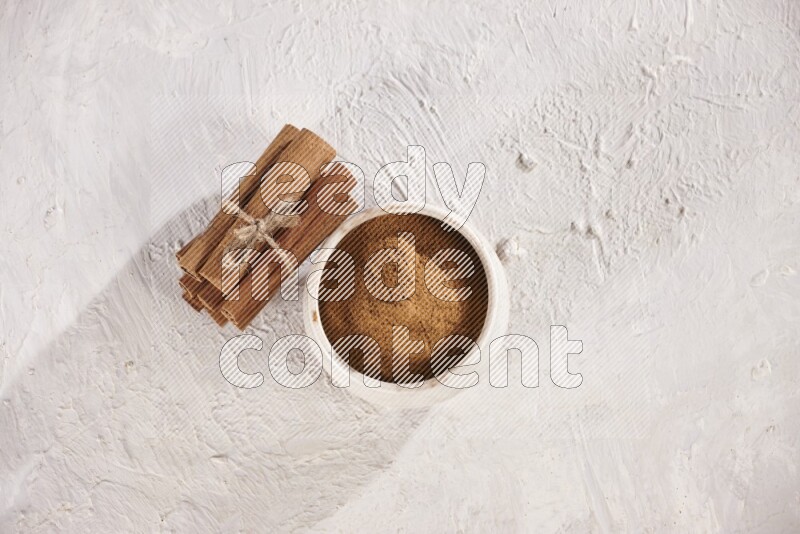 Cinnamon sticks stacked and bounded beside a beige bowl full of cinnamon powder on white background