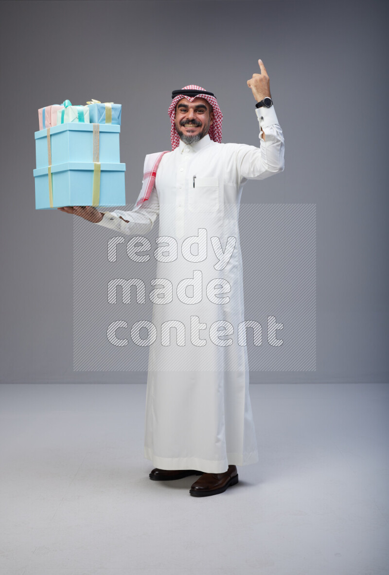 Saudi man Wearing Thob and red Shomag standing holding gift box on Gray background
