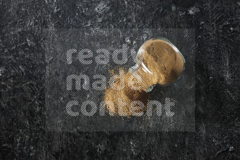 وعاء زجاجي مقلوب ممتلئ ببودرة الكمون والبودرة تناثرت منه على خلفية سوداء