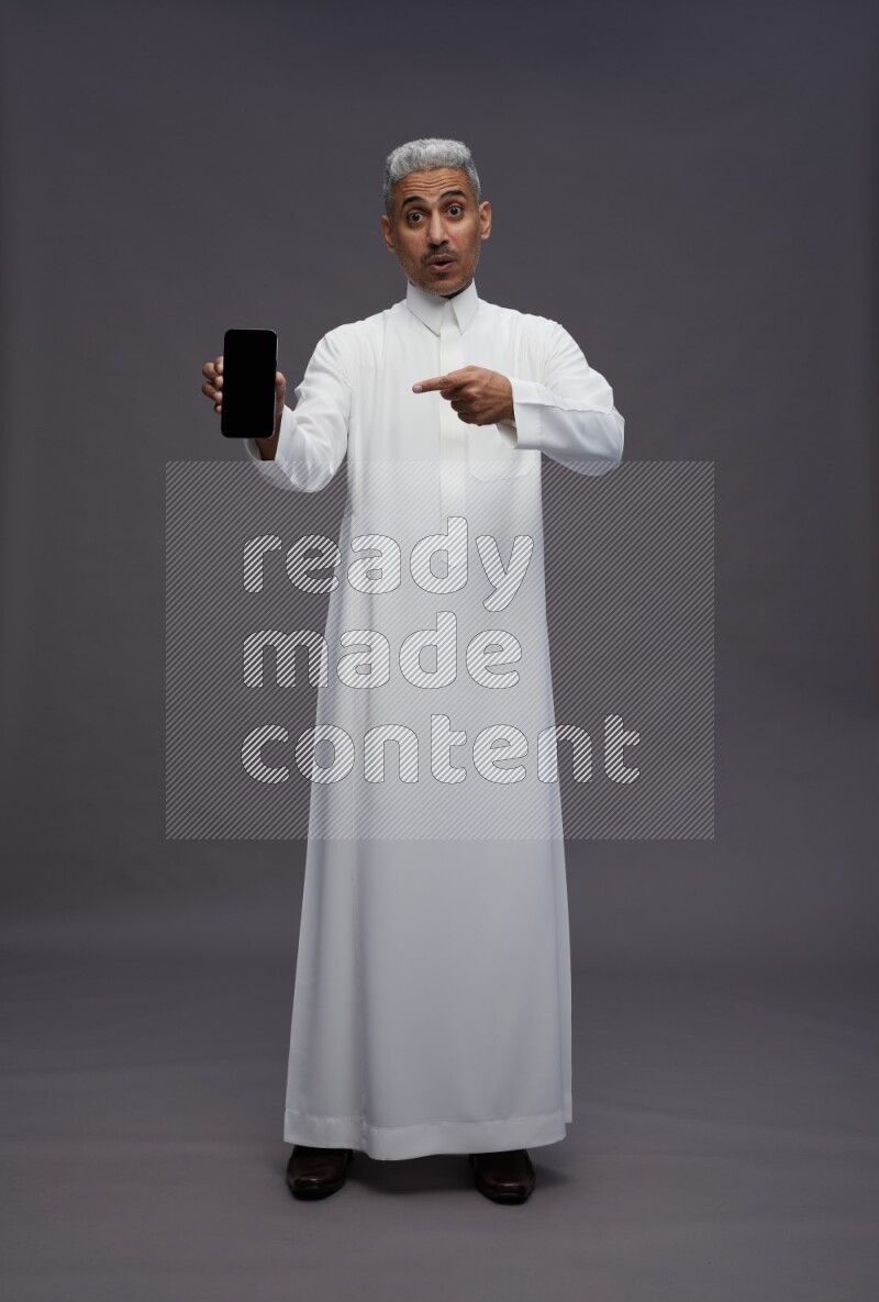 Saudi man wearing thob standing showing phone to camera on gray background