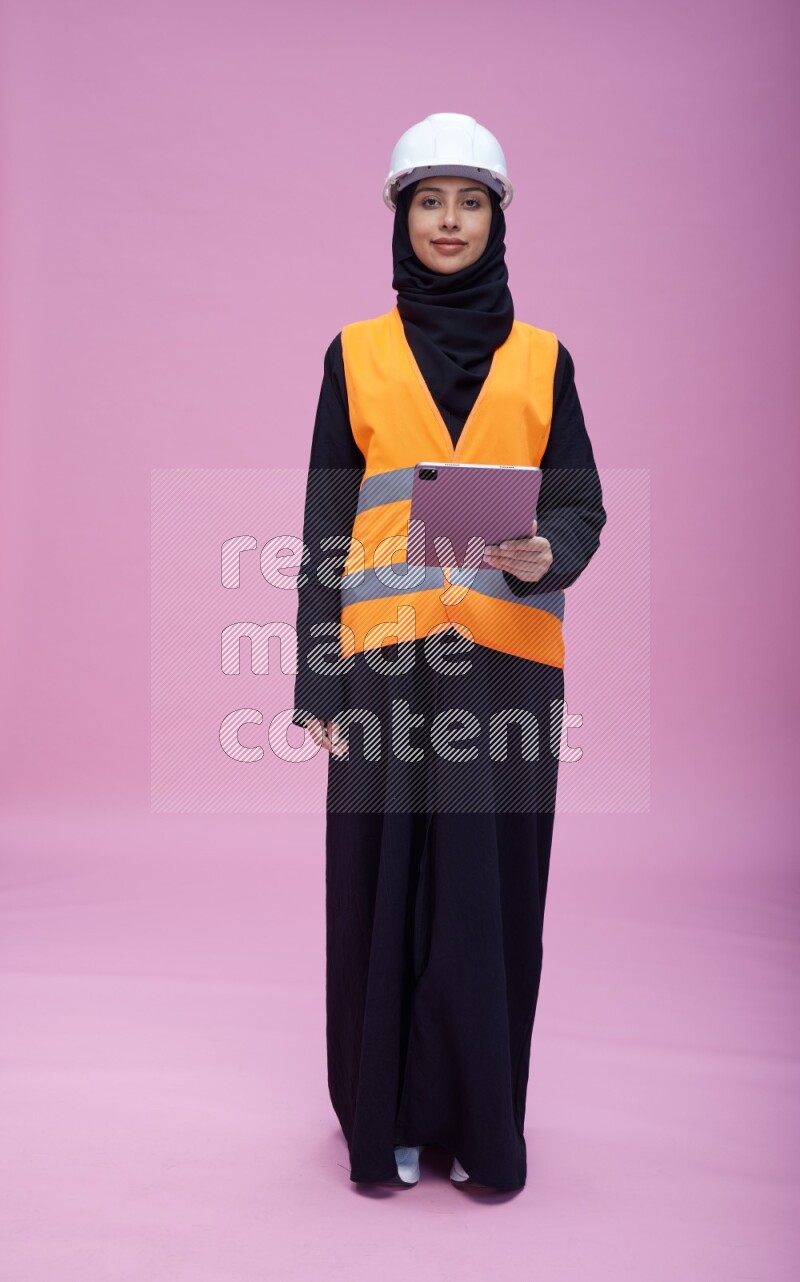 Saudi woman wearing Abaya with engineer vest and helmet standing working on tablet on pink background