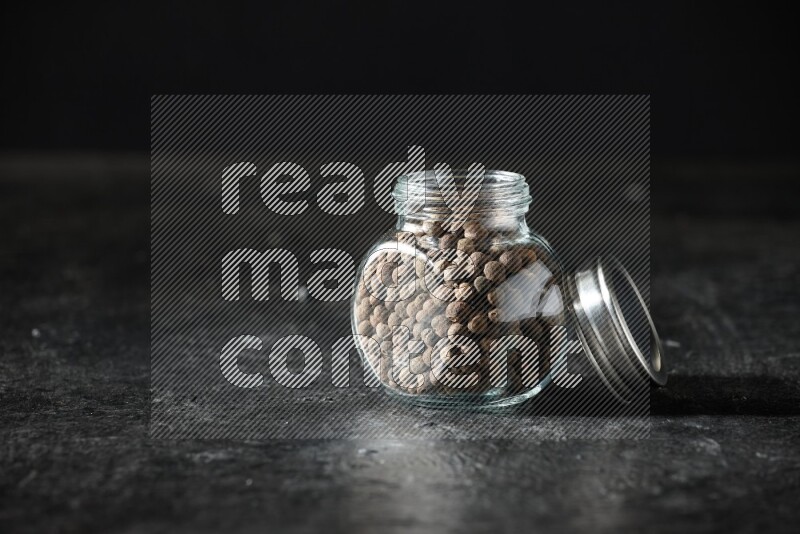 A glass spice jar full of allspice whole balls on a textured black flooring