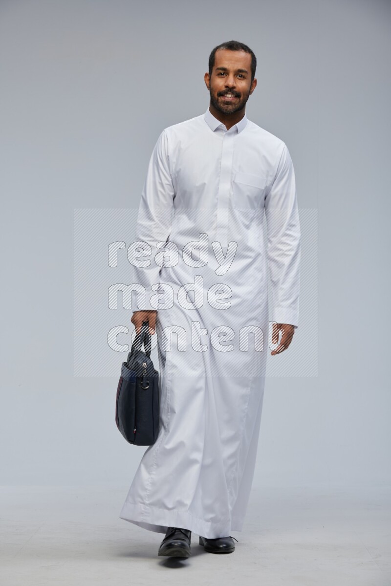Saudi man wearing thob standing holding suitcase on gray background