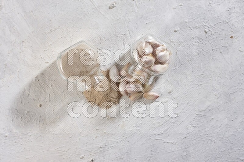 2 glass spice jars, one full of garlic powder and the other full of garlic cloves both flipped on a textured white flooring