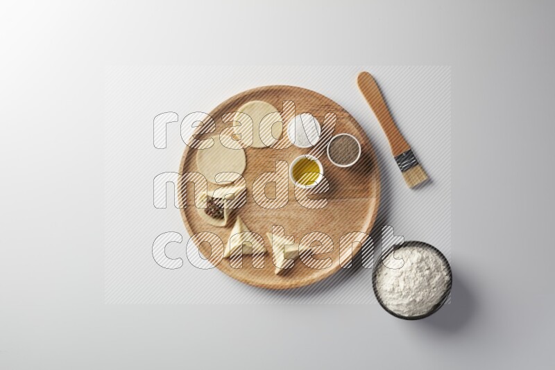 two closed sambosas and one open sambosa filled with meat while flour, salt, black pepper and oil with oil brush aside in a wooden dish on a white background