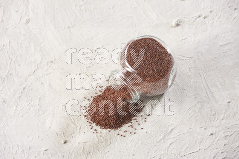 A glass spice jar full of garden cress seeds and jar is flipped with fallen seeds on a textured white flooring