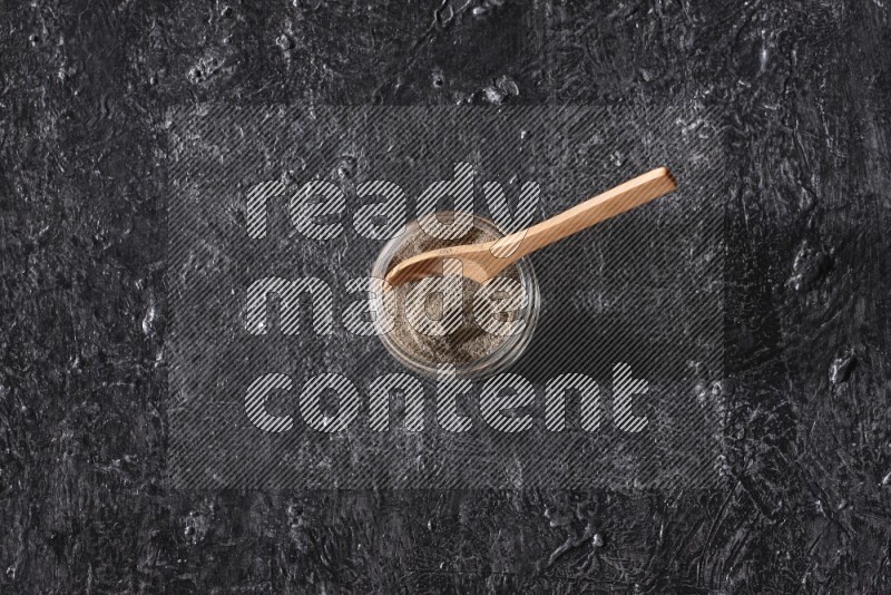 بودرة الفلفل الأسود في وعاء زجاجي مع ملعقة خشبية علي خلفية سوداء